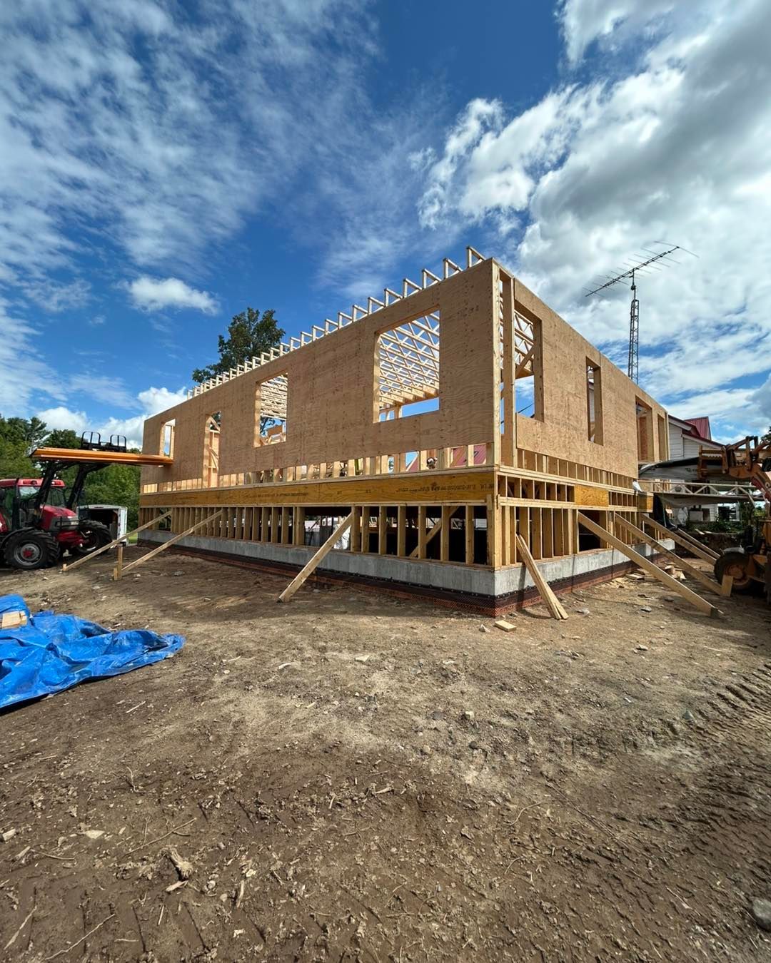 Chantier de construction, bâtiment à ossature bois en cours de construction, soutenu par des poteaux, sous un ciel bleu nuageux.