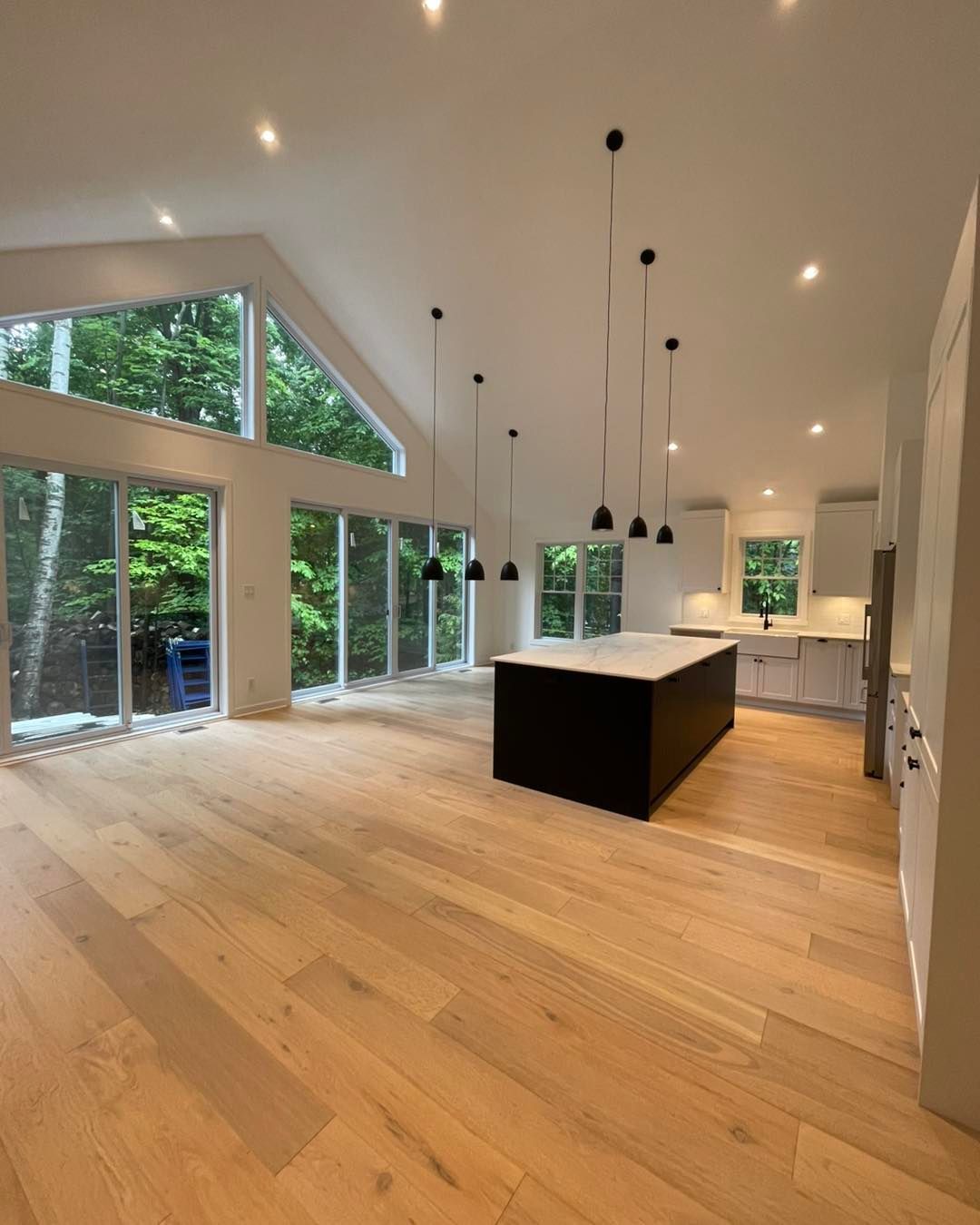 Cuisine moderne avec parquet, îlot central noir, placards blancs et grandes fenêtres donnant sur les arbres.