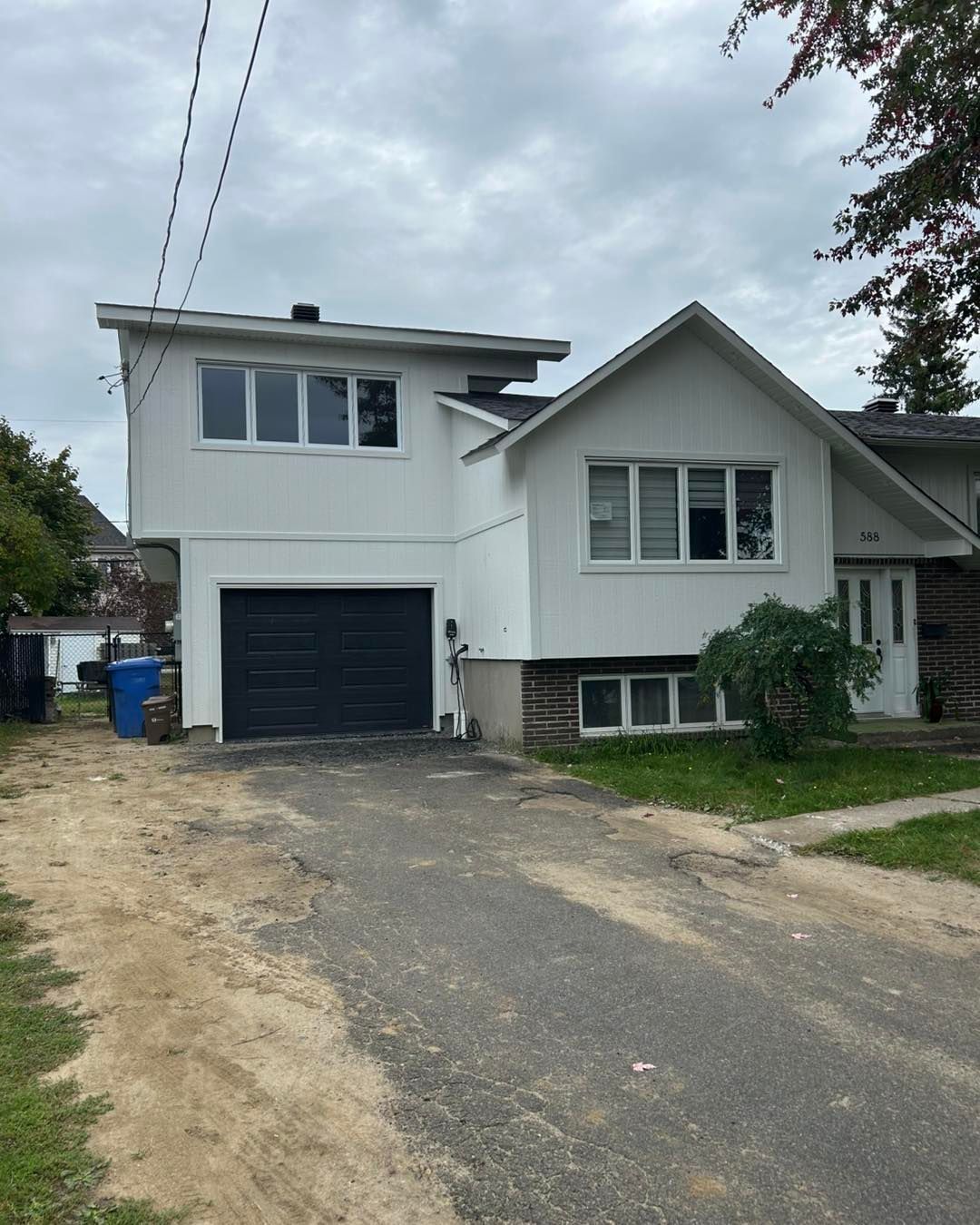 Maison blanche à deux étages avec porte de garage et allée noires, sous un ciel nuageux.