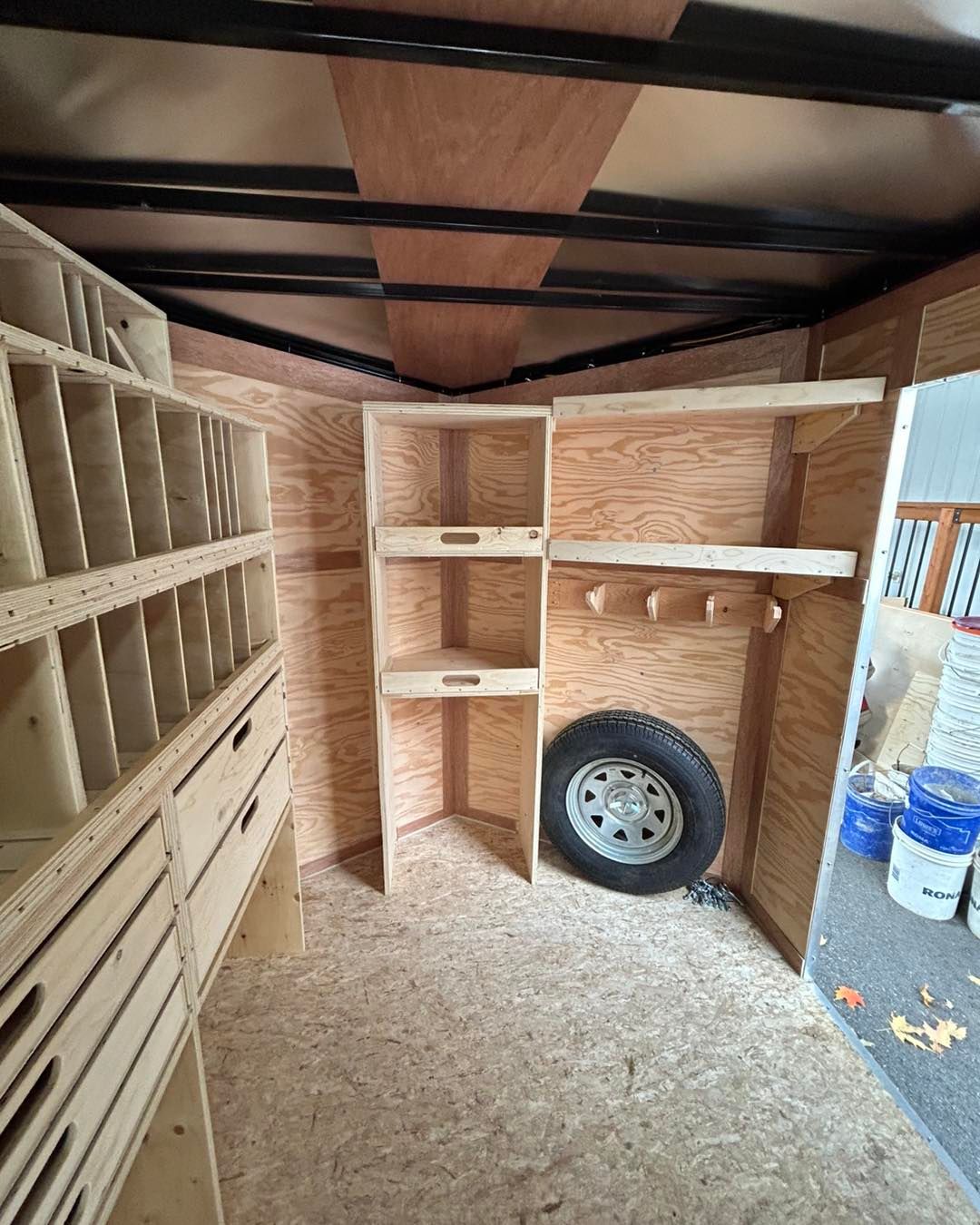 Intérieur d'une remorque cargo. Des étagères et des rangements en bois tapissent les parois. Roue de secours montée.