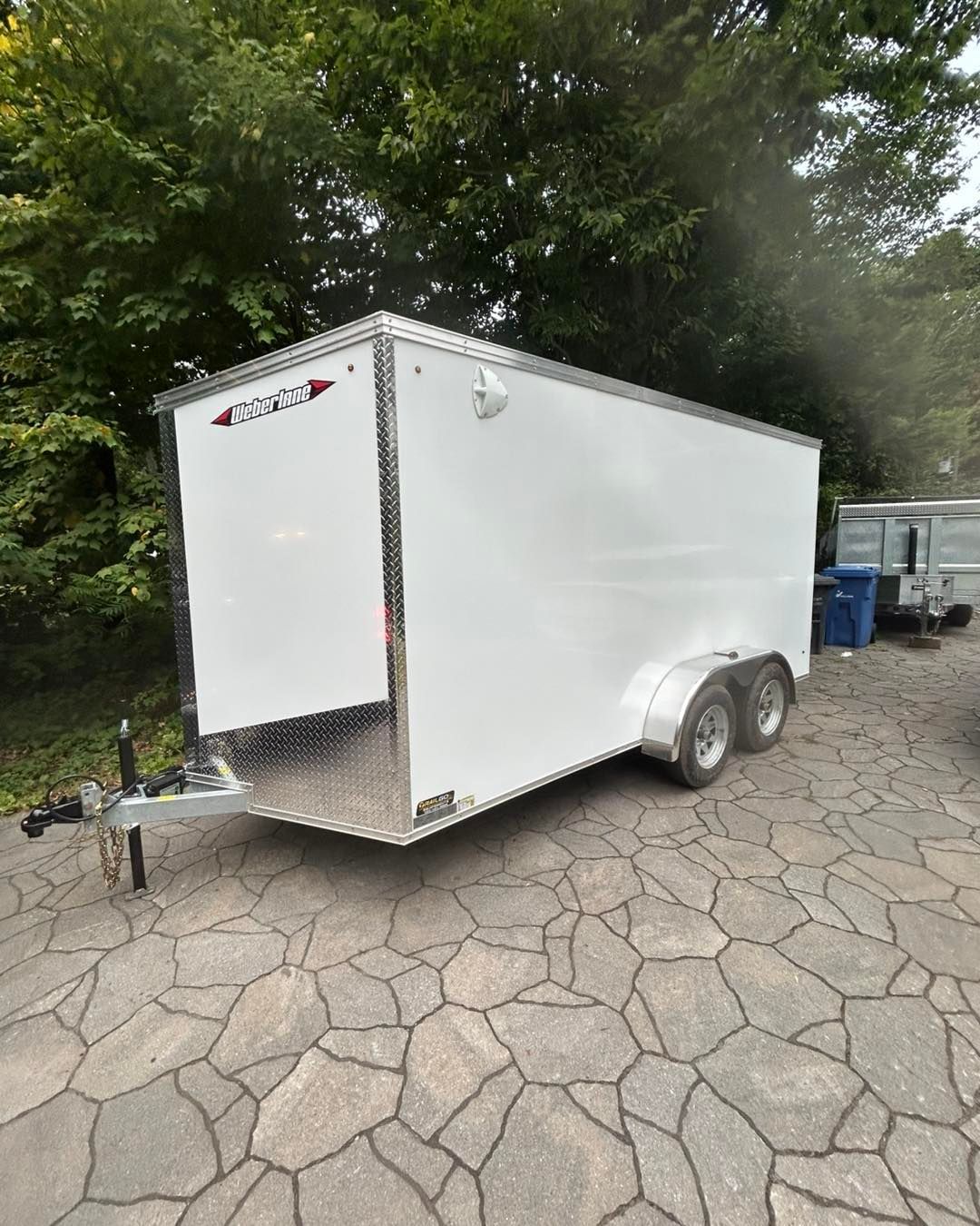 Remorque cargo blanche fermée sur une allée de pierre, adossée à des arbres.
