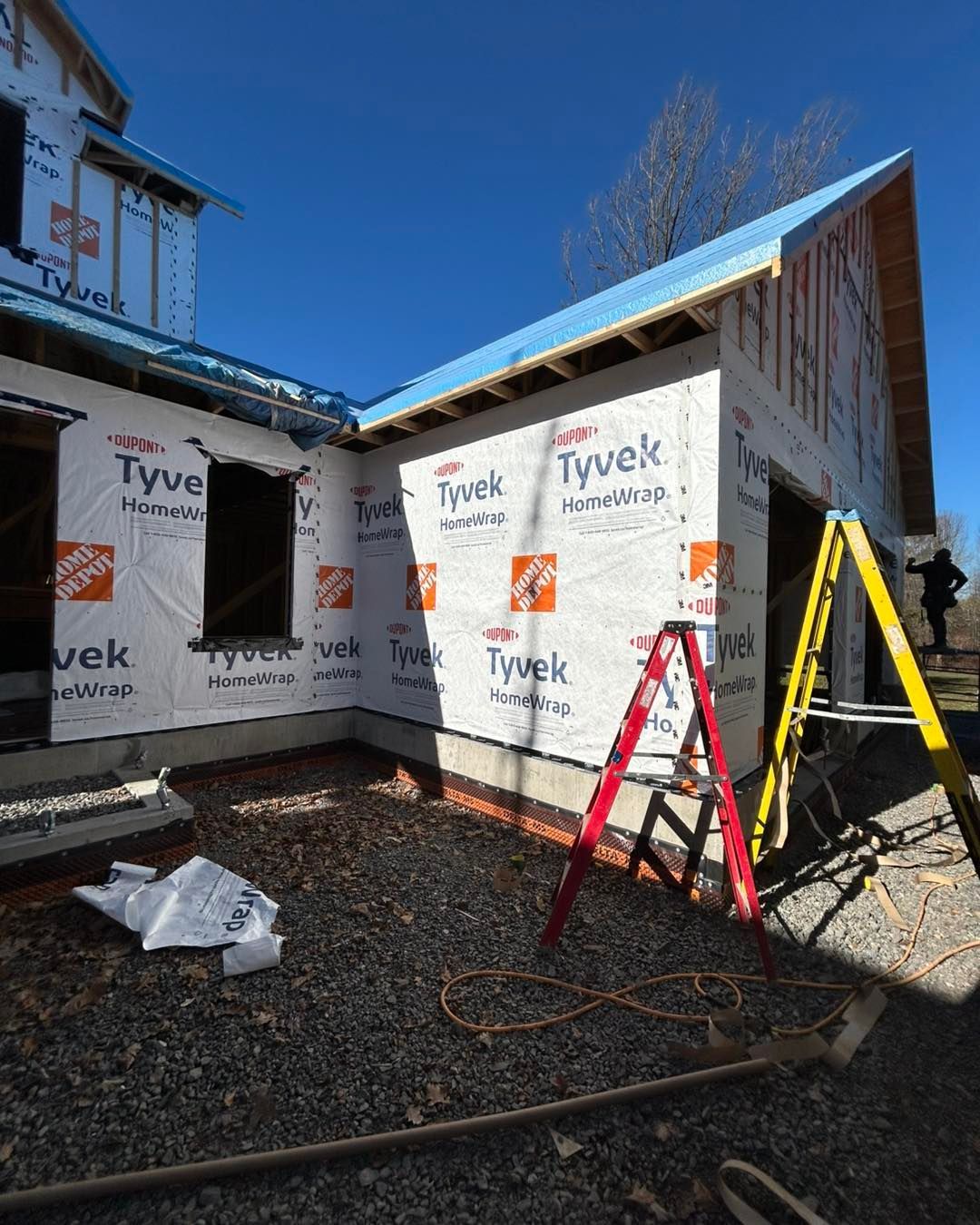 Bâtiment en construction, vue extérieure. La structure est recouverte d'une bâche Tyvek ; ciel bleu au-dessus. Échelle en A et échelle rouge visibles.