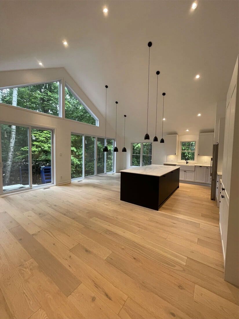 Cuisine moderne et spacieuse avec parquet, îlot central foncé, armoires blanches et grandes fenêtres donnant sur les arbres.