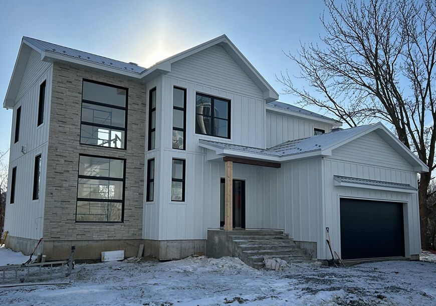 Maison blanche à deux étages avec fenêtres à cadre noir, façade en briques et garage, dans un décor enneigé.