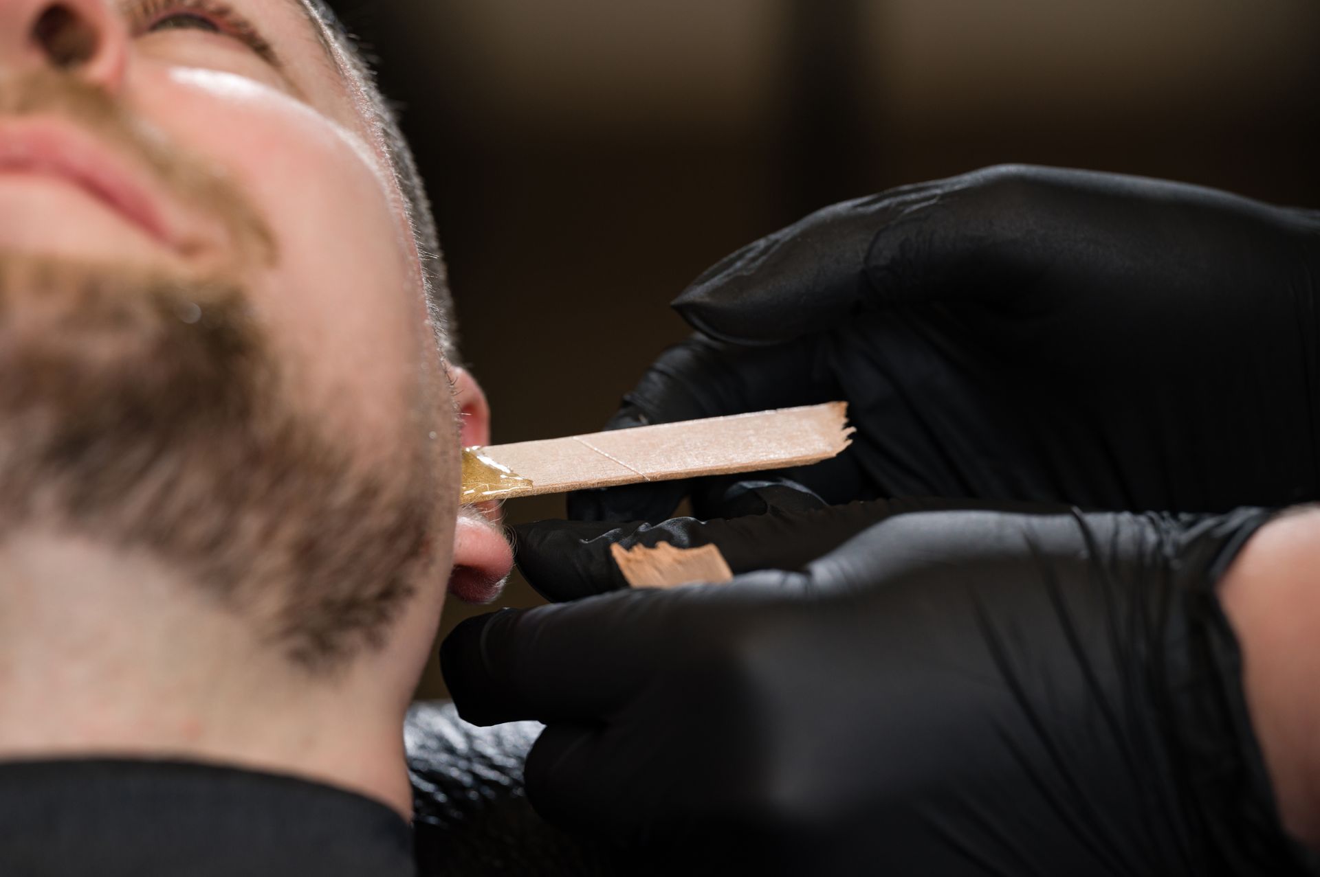 Person getting chest waxed; gloved hands apply black wax with a wooden stick.