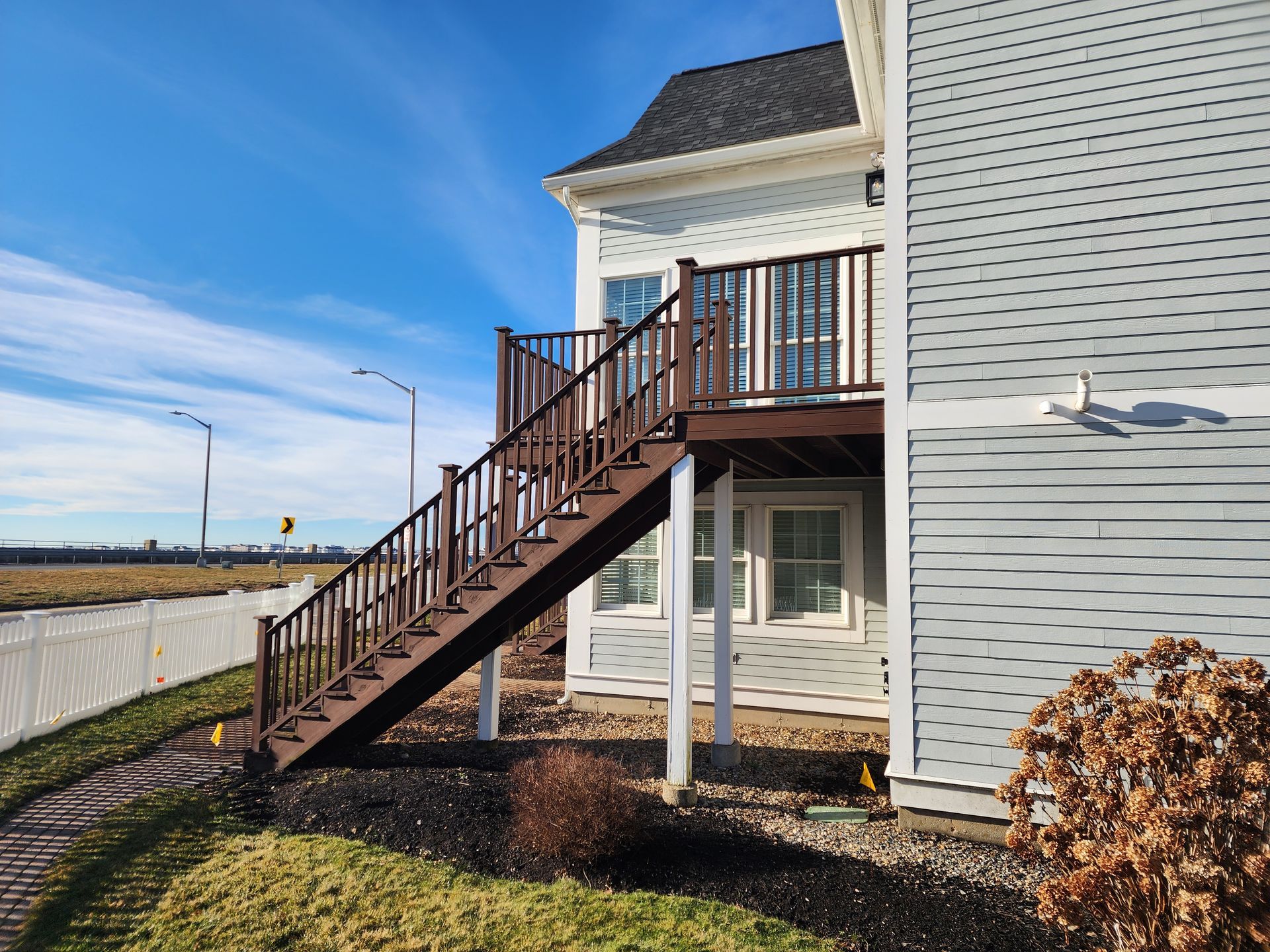 Brown staircase leading to a wooden deck on a two-story house with blue siding, under a clear, sunny sky.