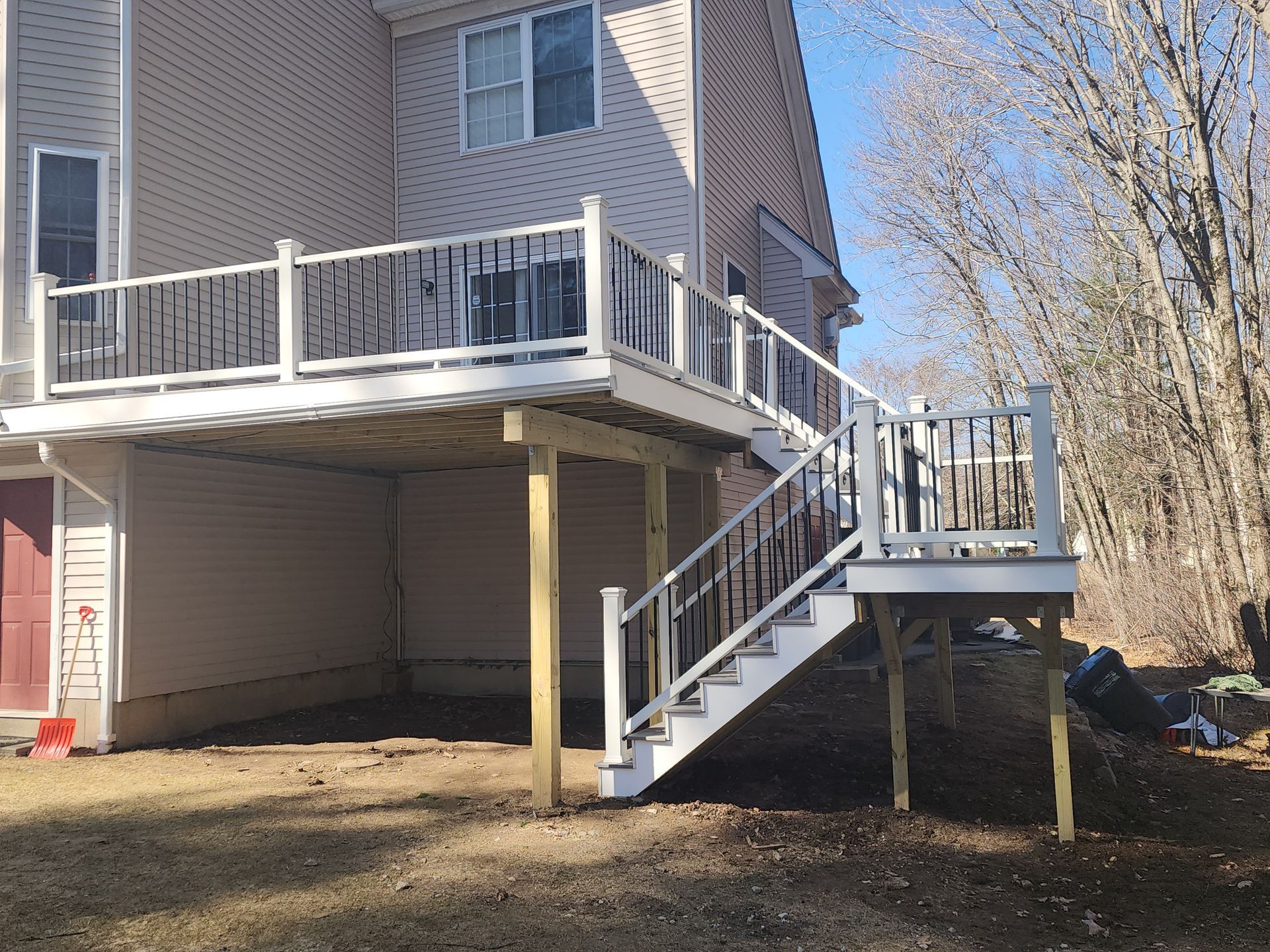 Backyard deck with stairs, two-story house, white railing, black spindles, wooden support beams, and brown backyard.