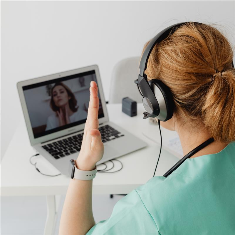 Medical professional in scrubs, headset, waves to person on laptop screen during a telehealth appointment.