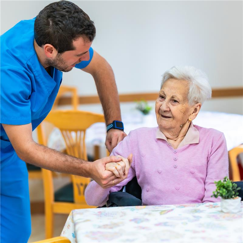 Caregiver in blue helping smiling elderly woman in pink sit at a table.