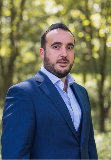 A man in a blue suit and white shirt is standing in front of trees.