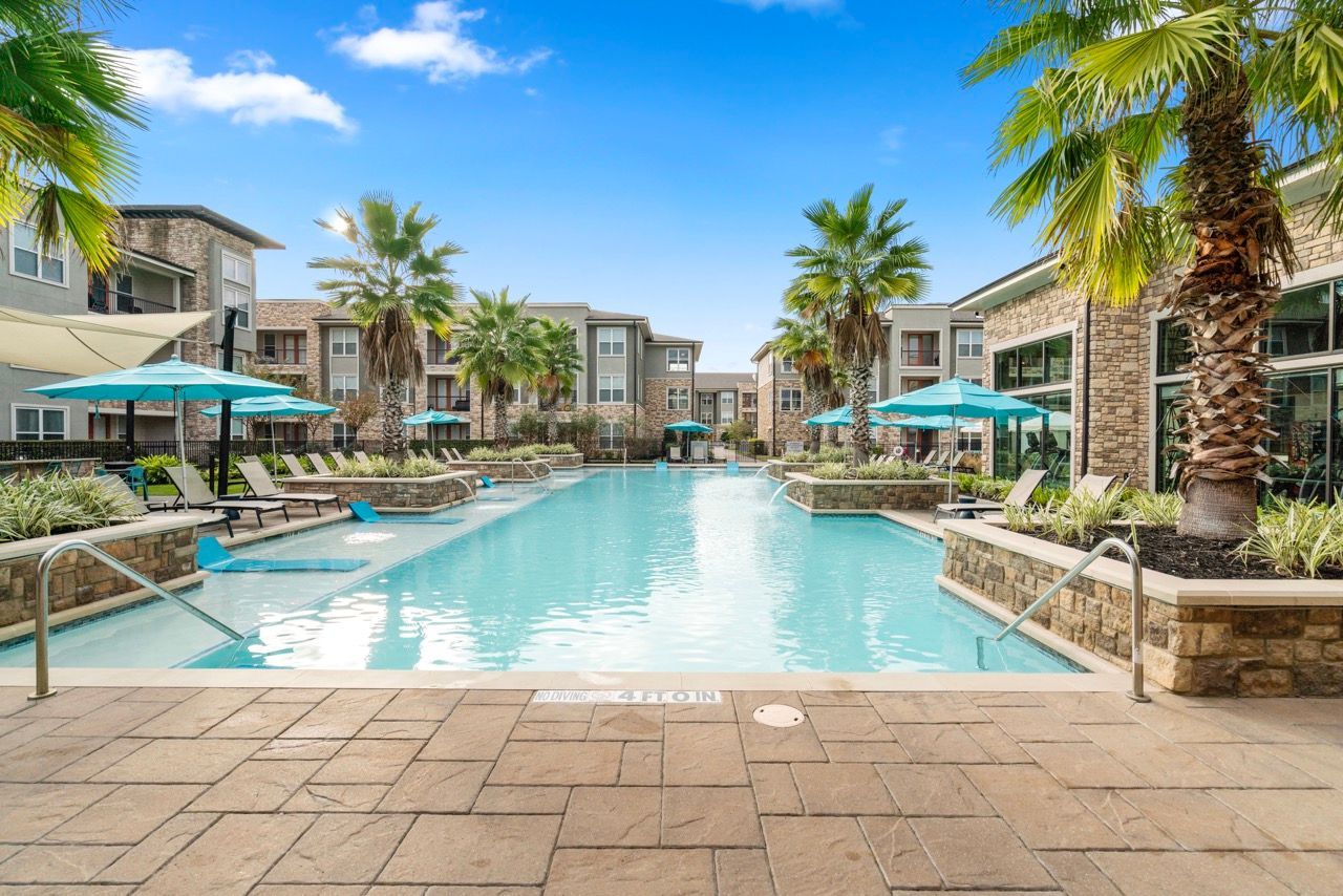 Resort-style apartment pool with palm trees, lounge chairs, and turquoise umbrellas.