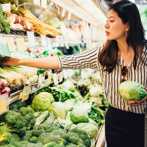 A person in a striped shirt shops for fresh produce, holding a head of lettuce while reaching for vegetables on a shelf.