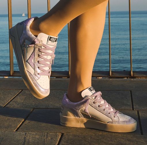 Donna che indossa scarpe da ginnastica bianche e color lavanda scintillanti, in piedi su una passerella vicino all'oceano.