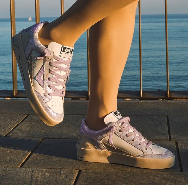 Donna che indossa scarpe da ginnastica bianche e color lavanda scintillanti, in piedi su una passerella vicino all'oceano.
