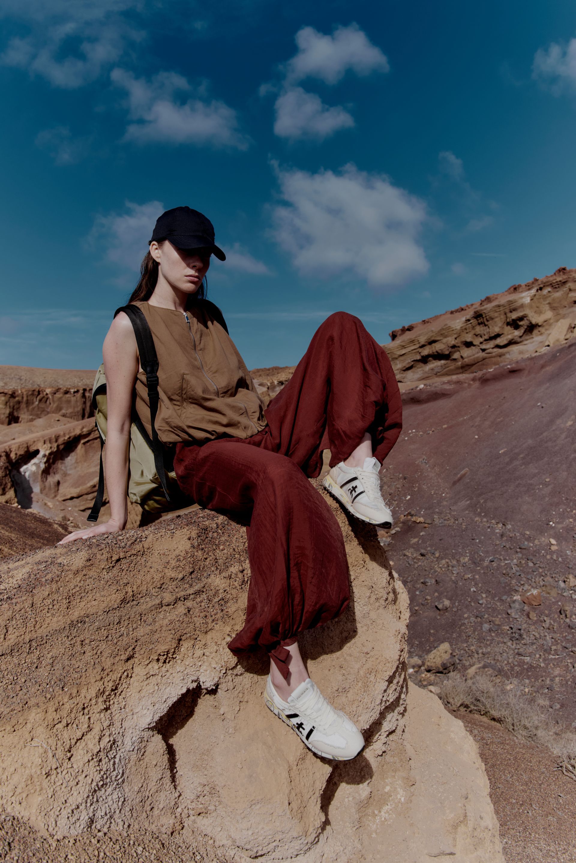 Persona accovacciata su un marciapiede con piumino, berretto e scarpe da ginnastica. Sullo sfondo città, giornata di sole.