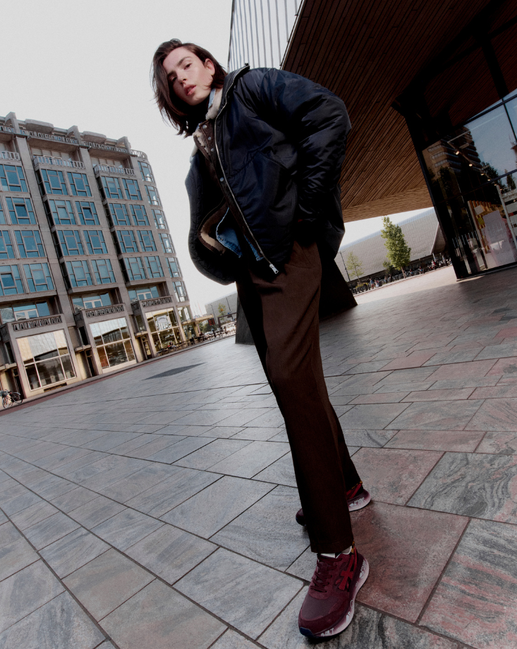 Uomo con giubbotto bomber e pantaloni marroni su un marciapiede urbano, con la scritta 