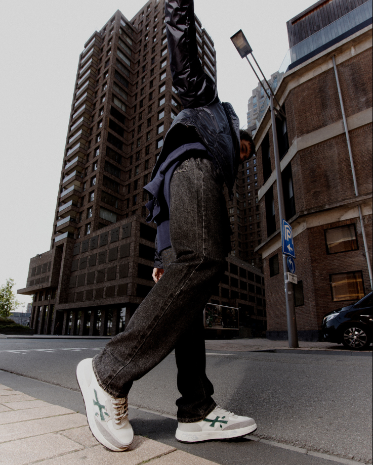 Persona in giacca nera e jeans, scarpe da ginnastica bianche, che cammina per strada con un cartello con scritto 