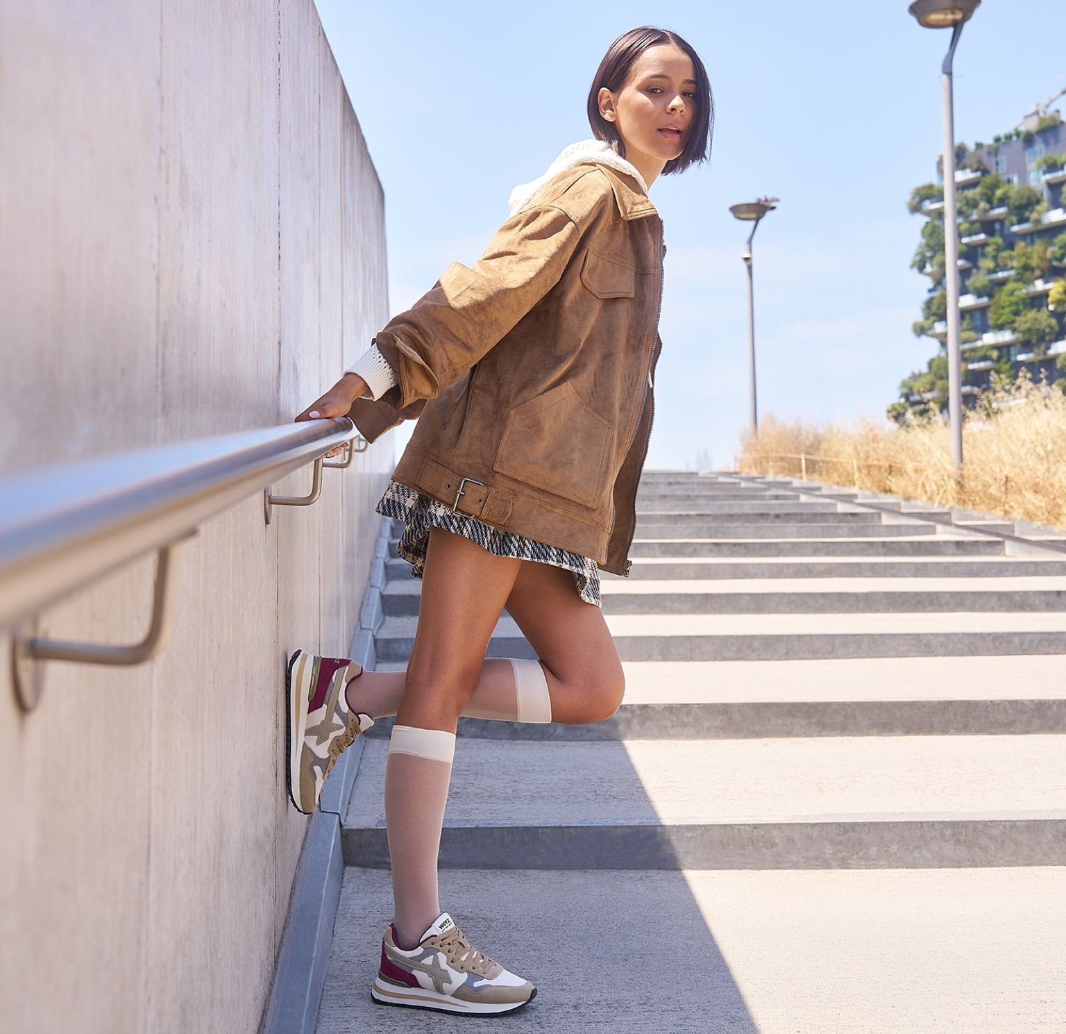 Uomo con giubbotto bomber e pantaloni marroni su un marciapiede urbano, con la scritta 