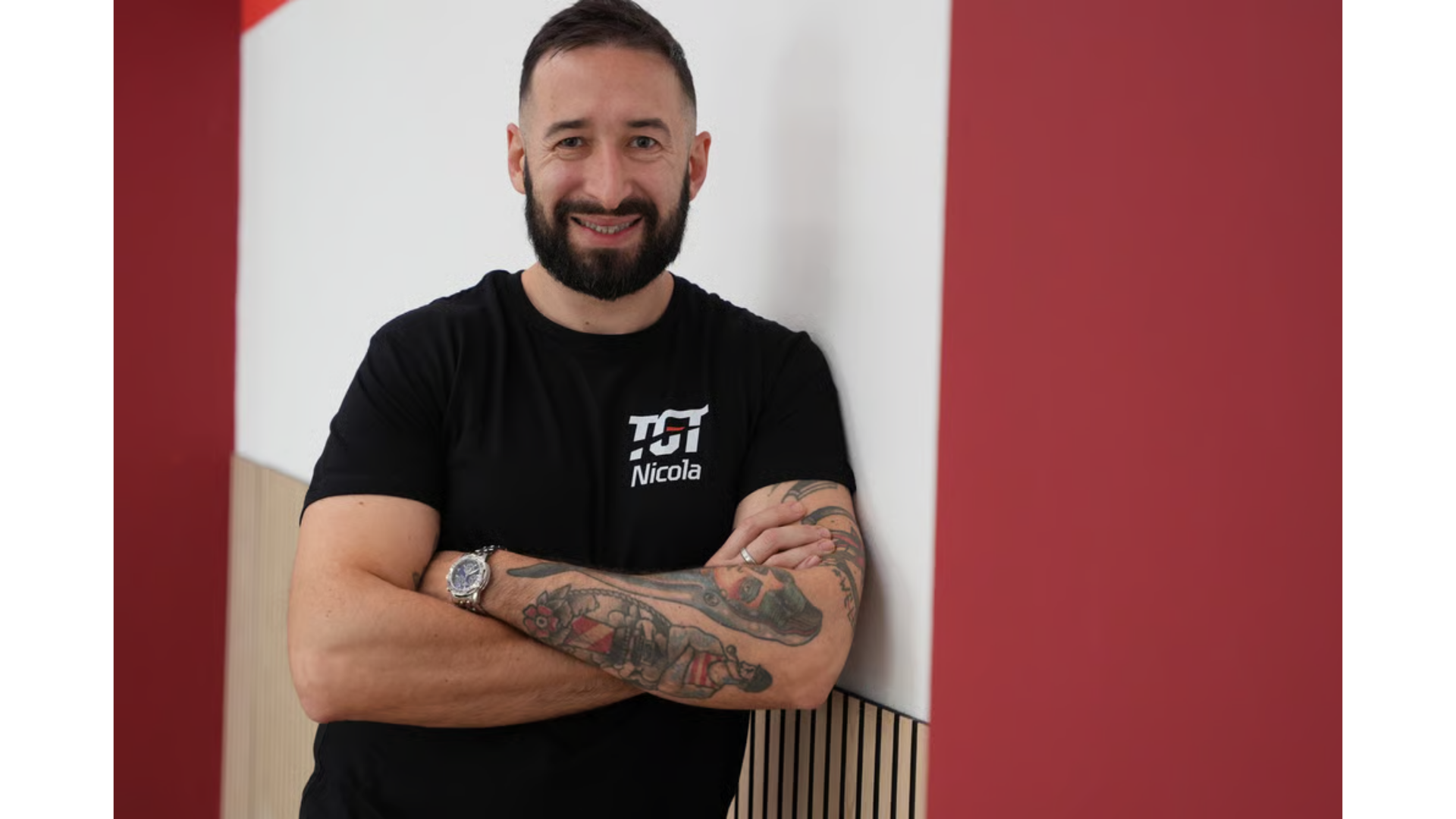 Una persona sorridente  una maglietta nera posa a braccia incrociate contro un muro bianco e rosso.