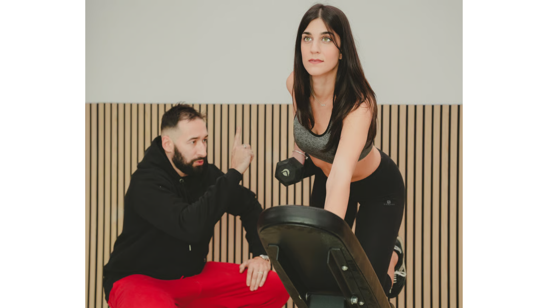 Un istruttore di fitness con felpa nera e pantaloni rossi dà istruzioni a una persona mentre solleva un manubrio 