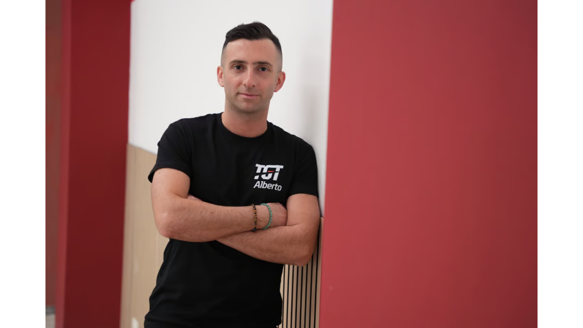 Una persona con indosso una maglietta nera, a braccia conserte, appoggiata a un muro bianco con una colonna rossa.