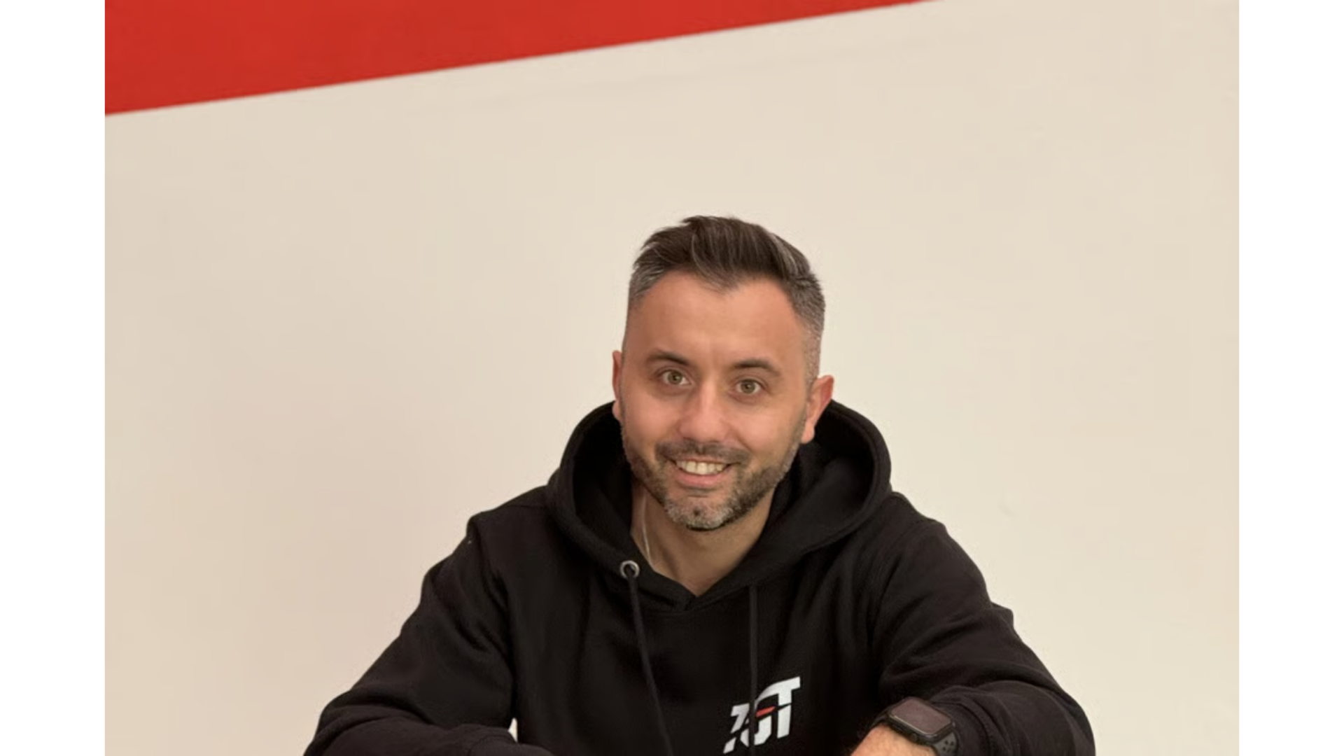 Una persona sorridente con indosso una felpa nera con cappuccio è seduta davanti a un muro bianco con una striscia rossa.