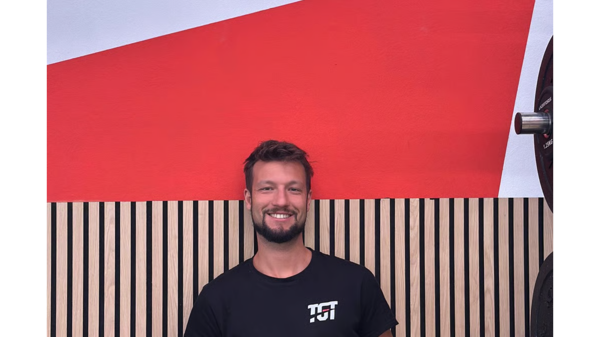 Una persona sorridente con la barba è in piedi davanti a uno sfondo caratterizzato da una parete rossa 