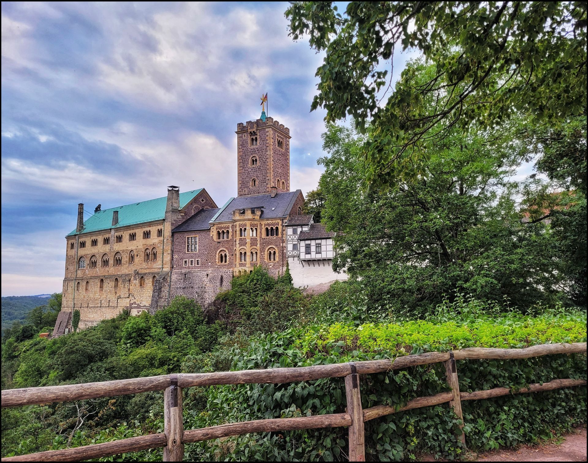 Mittelalterliche Wartburg auf einem Hügel, mit hohem Turm. Grünes Laubwerk umrahmt einen Holzzaun. Bewölkter Himmel.
