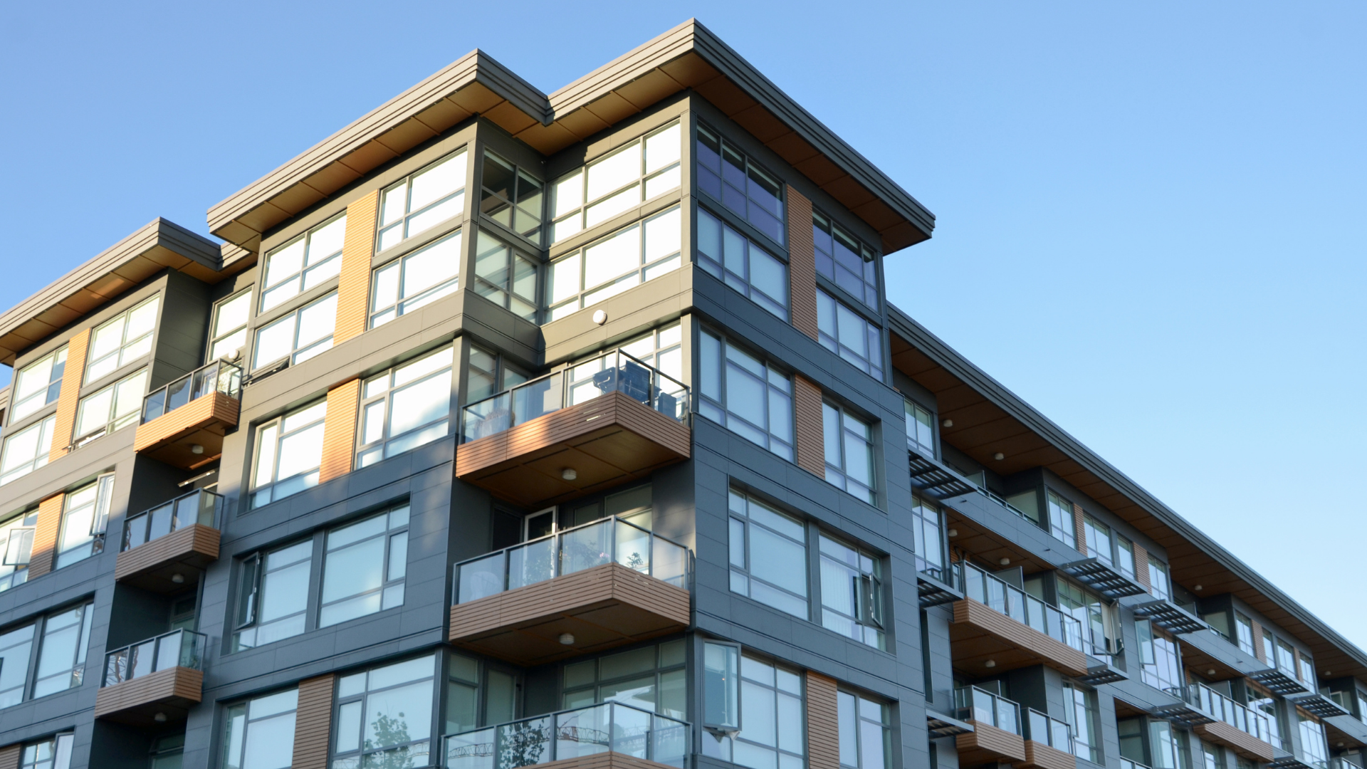 Modern multi-story apartment building with grey siding, wood accents, and large windows against a blue sky.