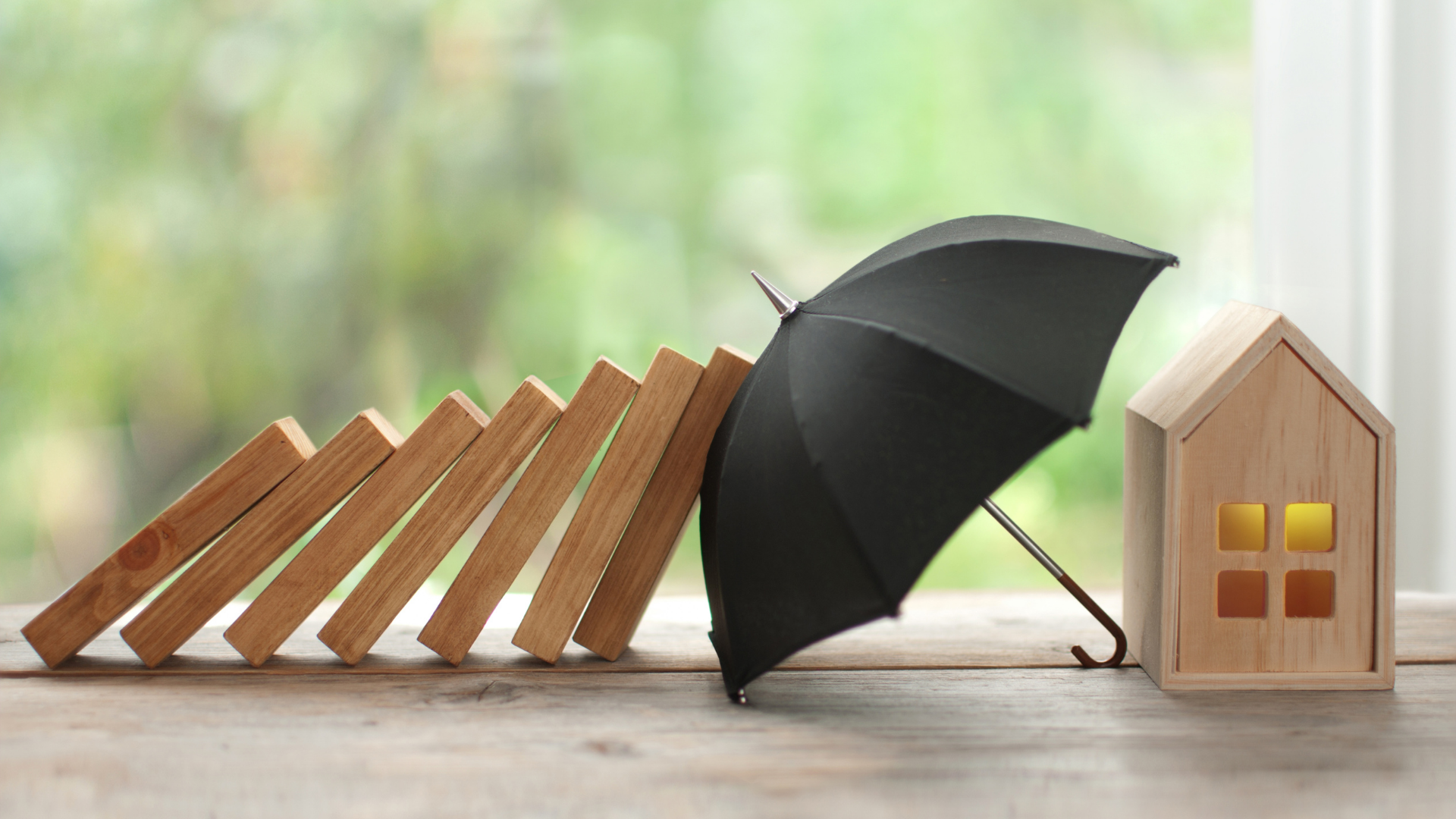 Wooden dominoes falling toward a toy house; black umbrella deflects them, on a table near a window.