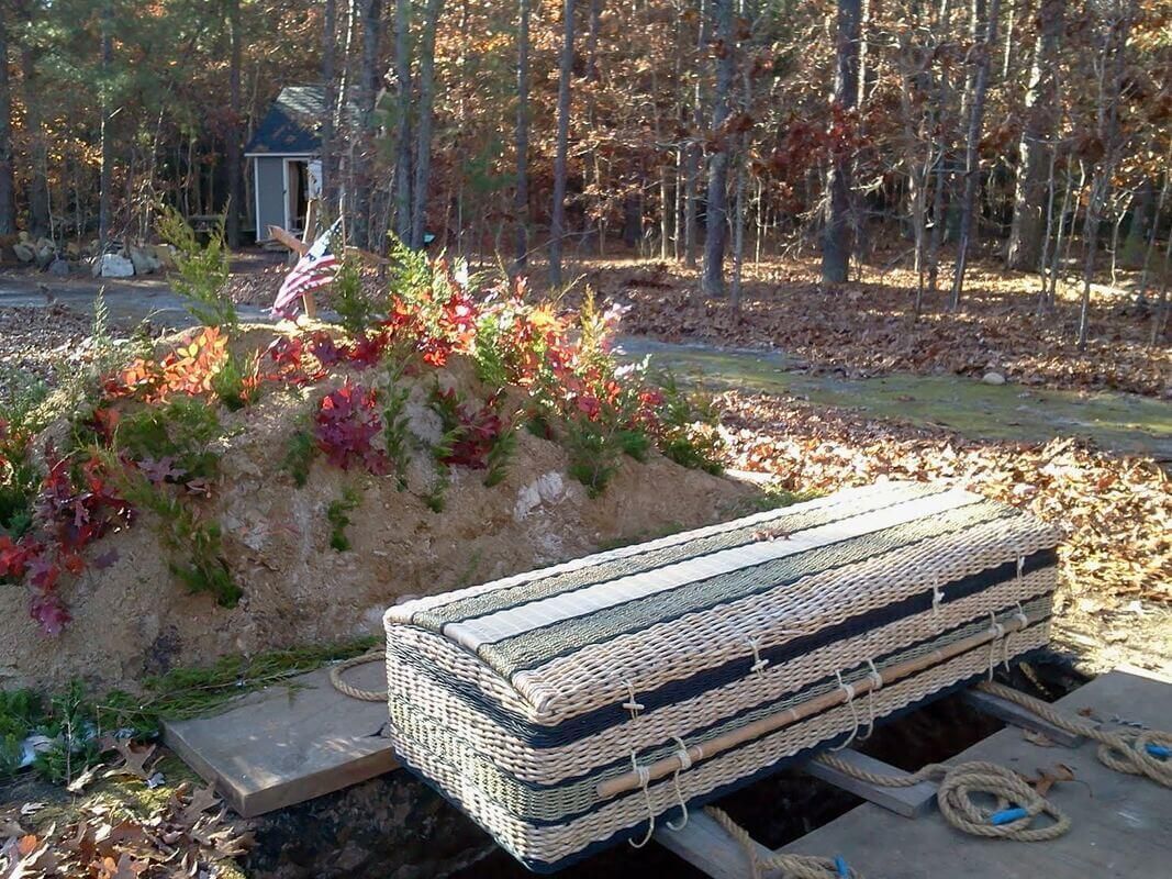 A casket resting over a grave, covered with rope. Flowers and foliage on a mound behind. Outdoors.