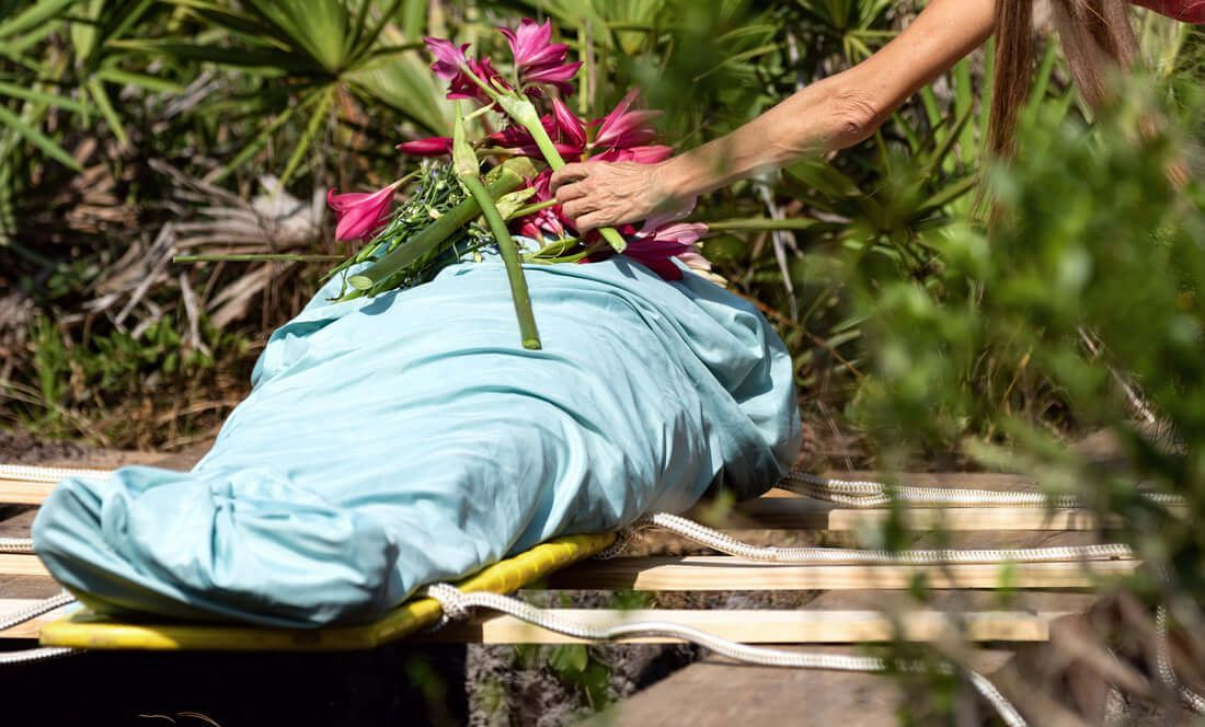 Person placing flowers on a body covered in a light blue shroud, outdoors.