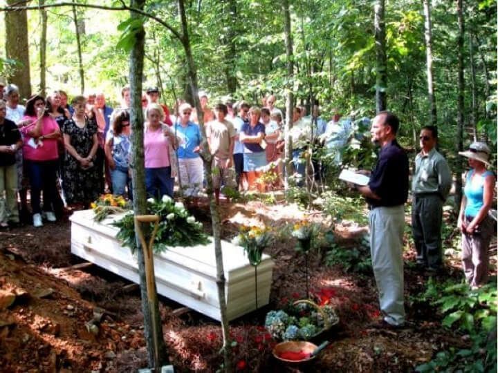 Funeral service in a wooded area; mourners surround a casket, a person is speaking.