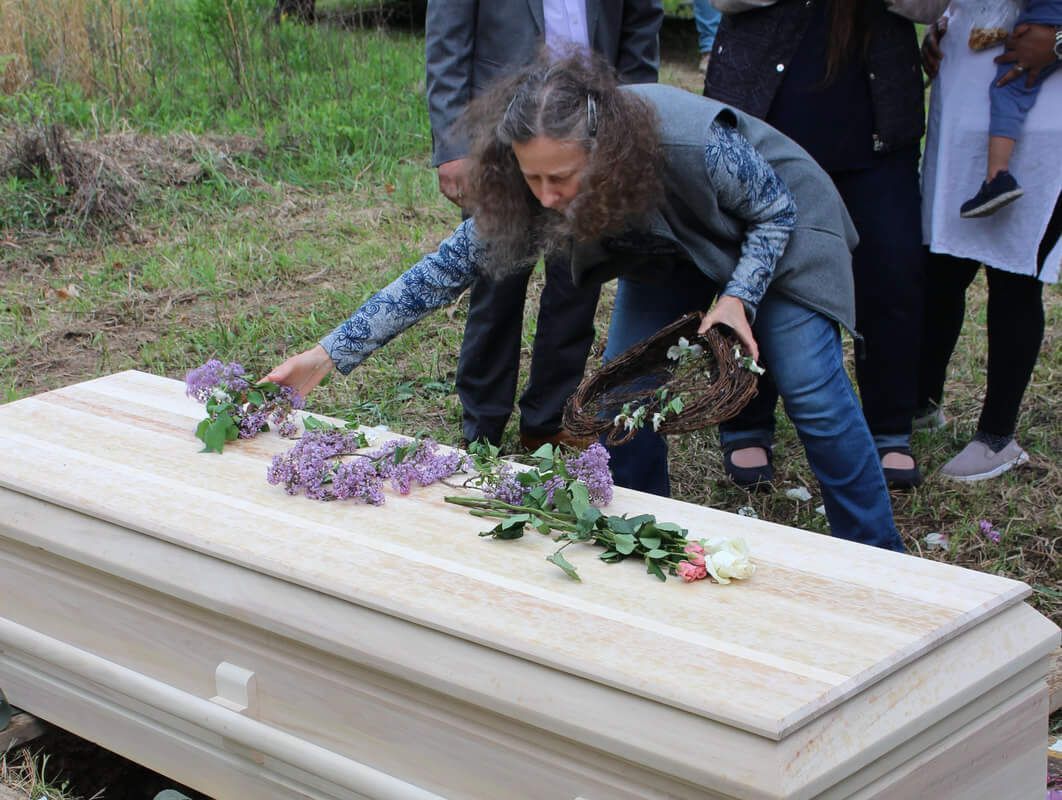 Person placing flowers on a casket outdoors. Other people are standing nearby, some holding a child.