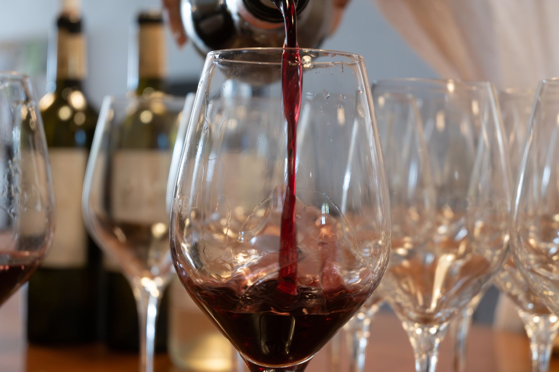 Vino rosso versato in un bicchiere, sullo sfondo altri bicchieri e bottiglie di vino.