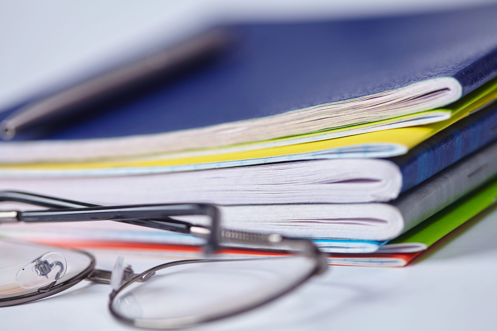 Pila di quaderni colorati con una penna sopra e degli occhiali in primo piano.