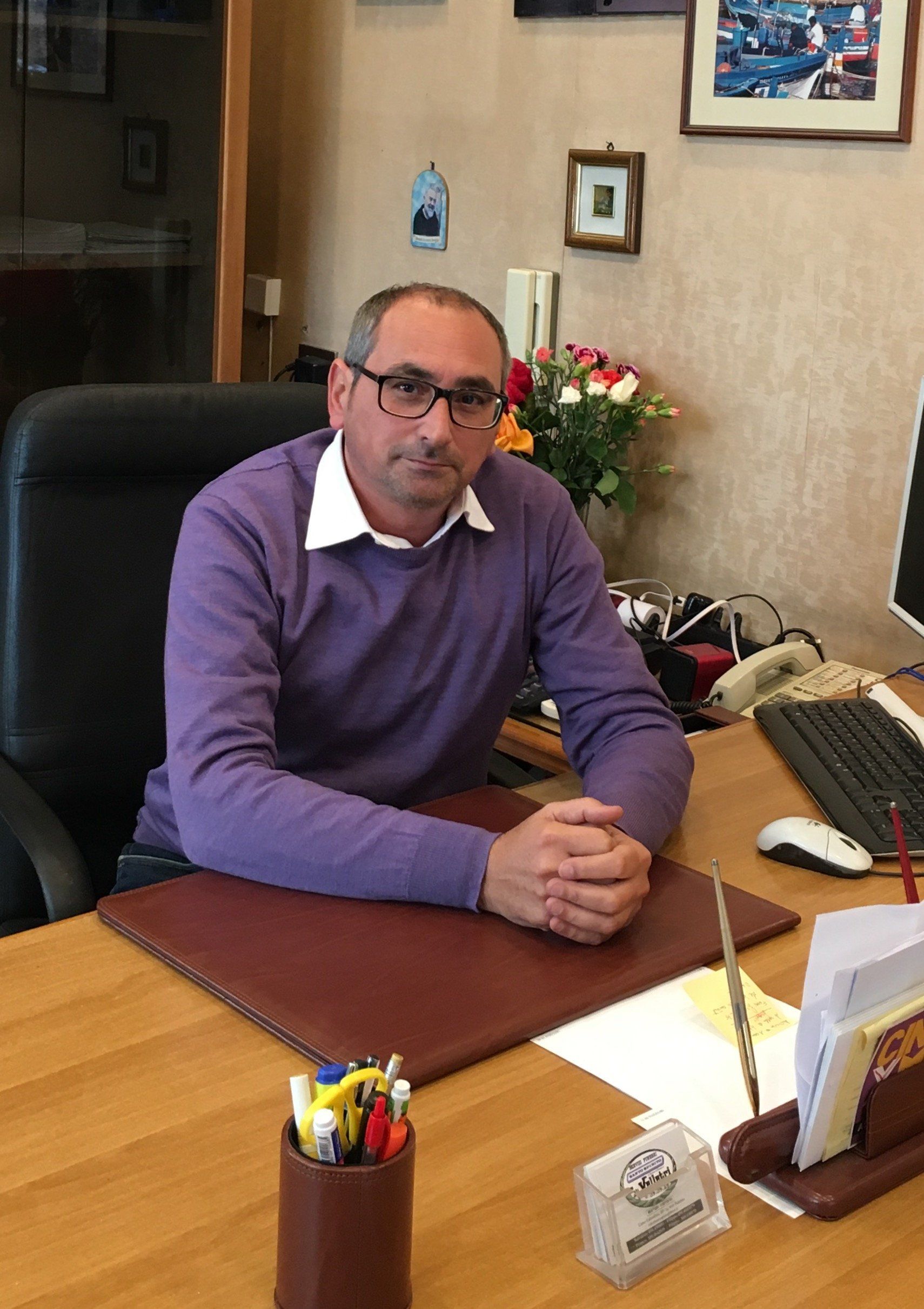 Un uomo con un maglione viola è seduto alla scrivania con le mani giunte e lo sguardo rivolto in avanti. Ambiente d'ufficio.