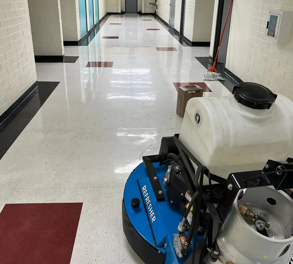 Blue floor scrubber machine in a bright hallway with white tile floors and red mats