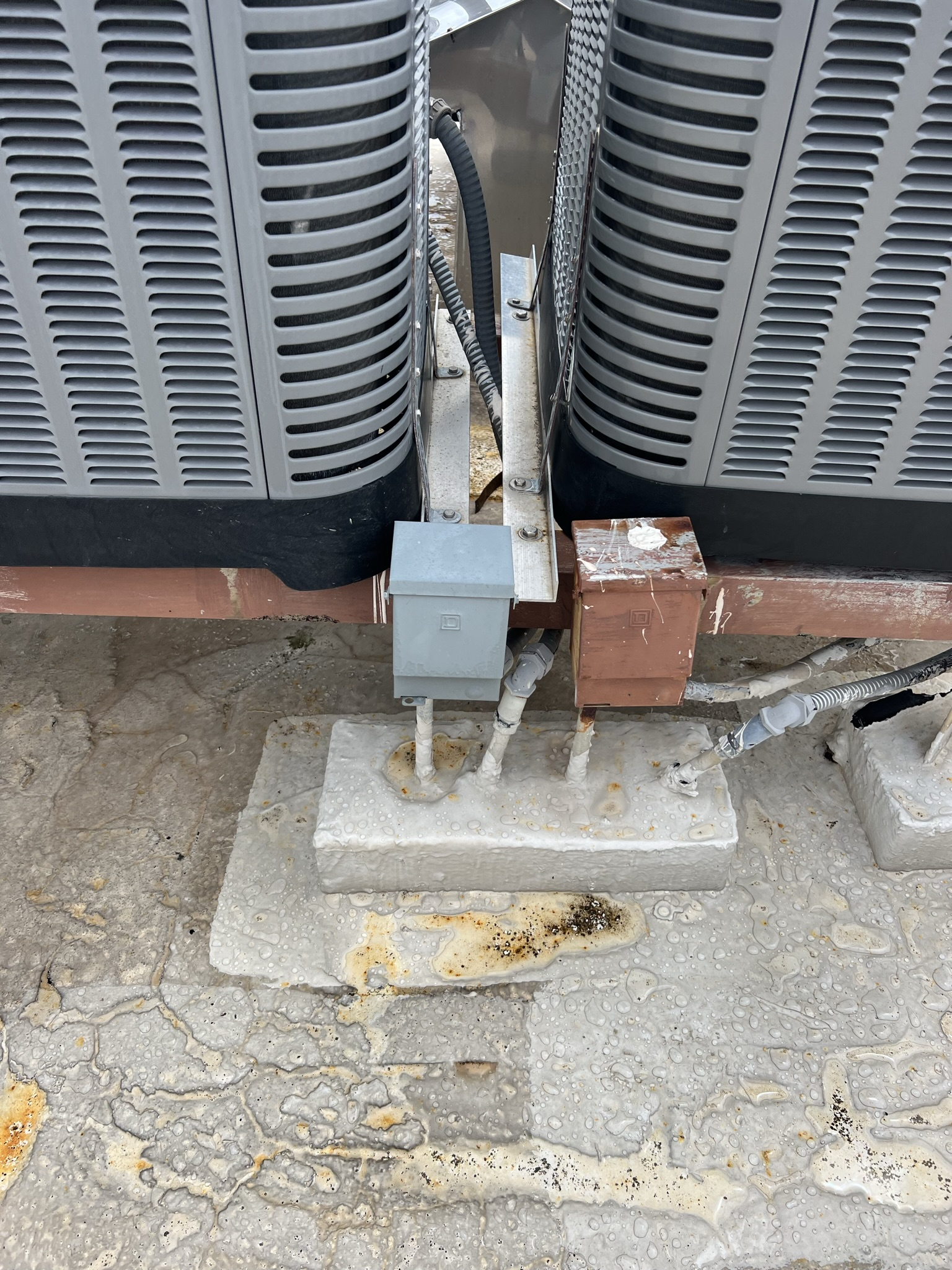 HVAC units on a rooftop, with electrical boxes and supports attached to a concrete base.