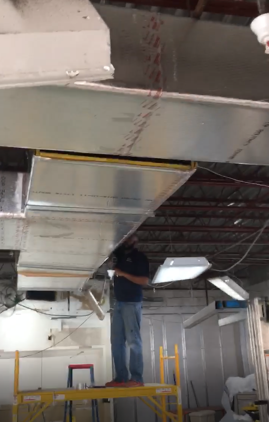 Man on scaffolding installing HVAC ductwork in a workshop, using tools.