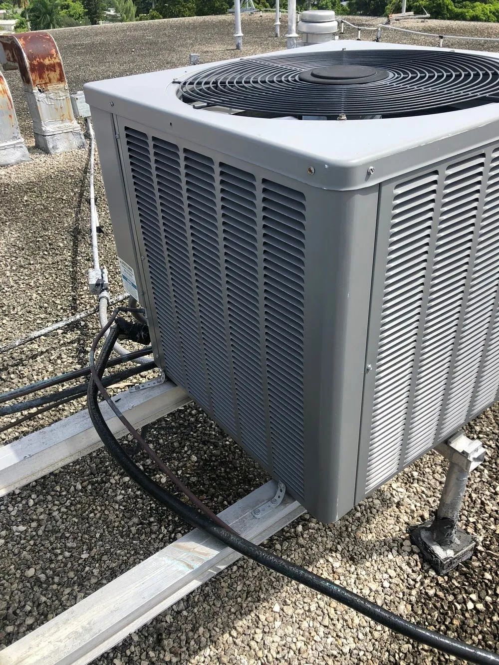 Outdoor air conditioning unit on a gravel roof, with black fan blades and gray metal housing.