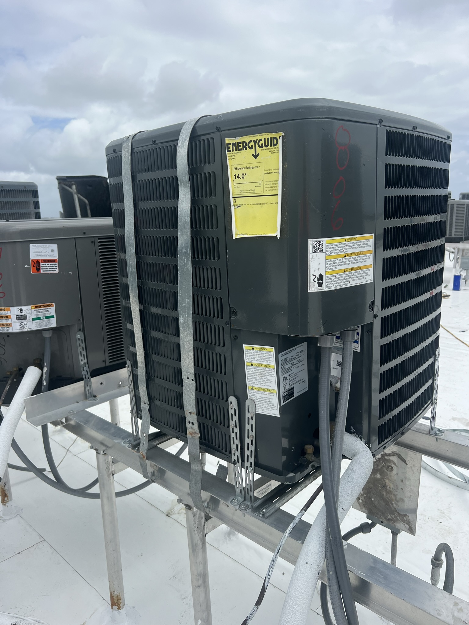 Black rooftop air conditioning unit on a white roof, secured with a metal strap.