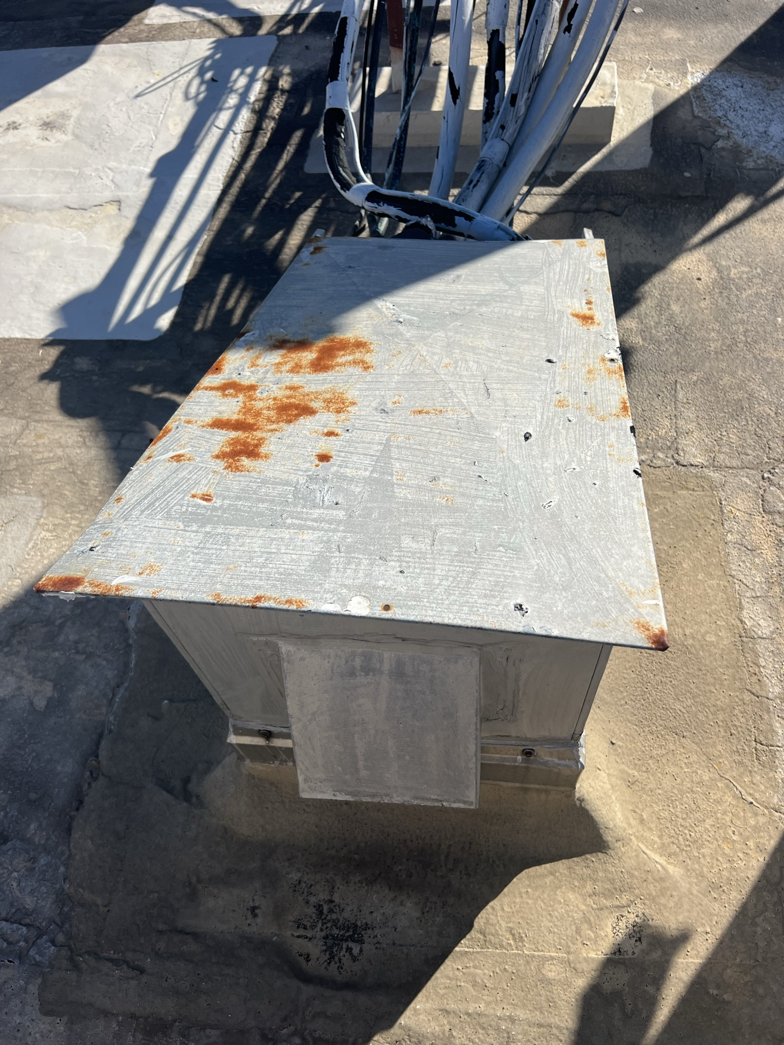 Rust-spotted metal box on concrete surface, beneath a structure with metal bars.