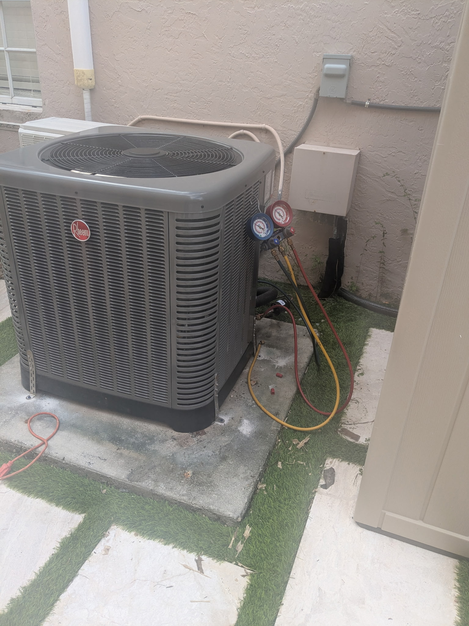 Air conditioning unit with gauges connected, set on concrete pavers, near a beige wall and faux grass.
