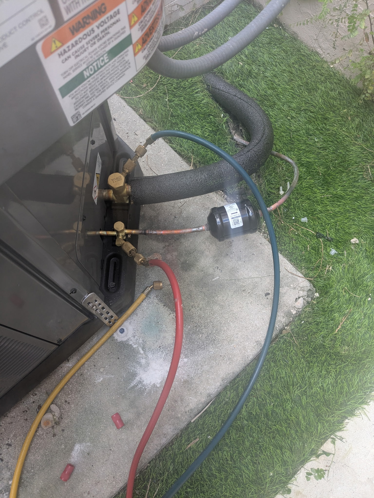 AC unit with red/blue hoses attached, outdoors on concrete, artificial grass nearby.