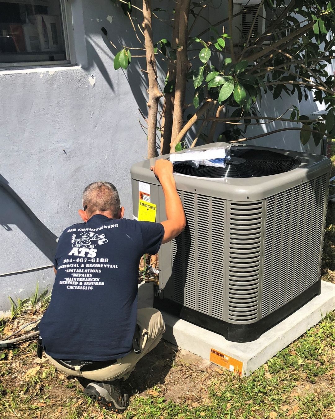 Man in blue shirt examines a gray air conditioning unit outdoors.