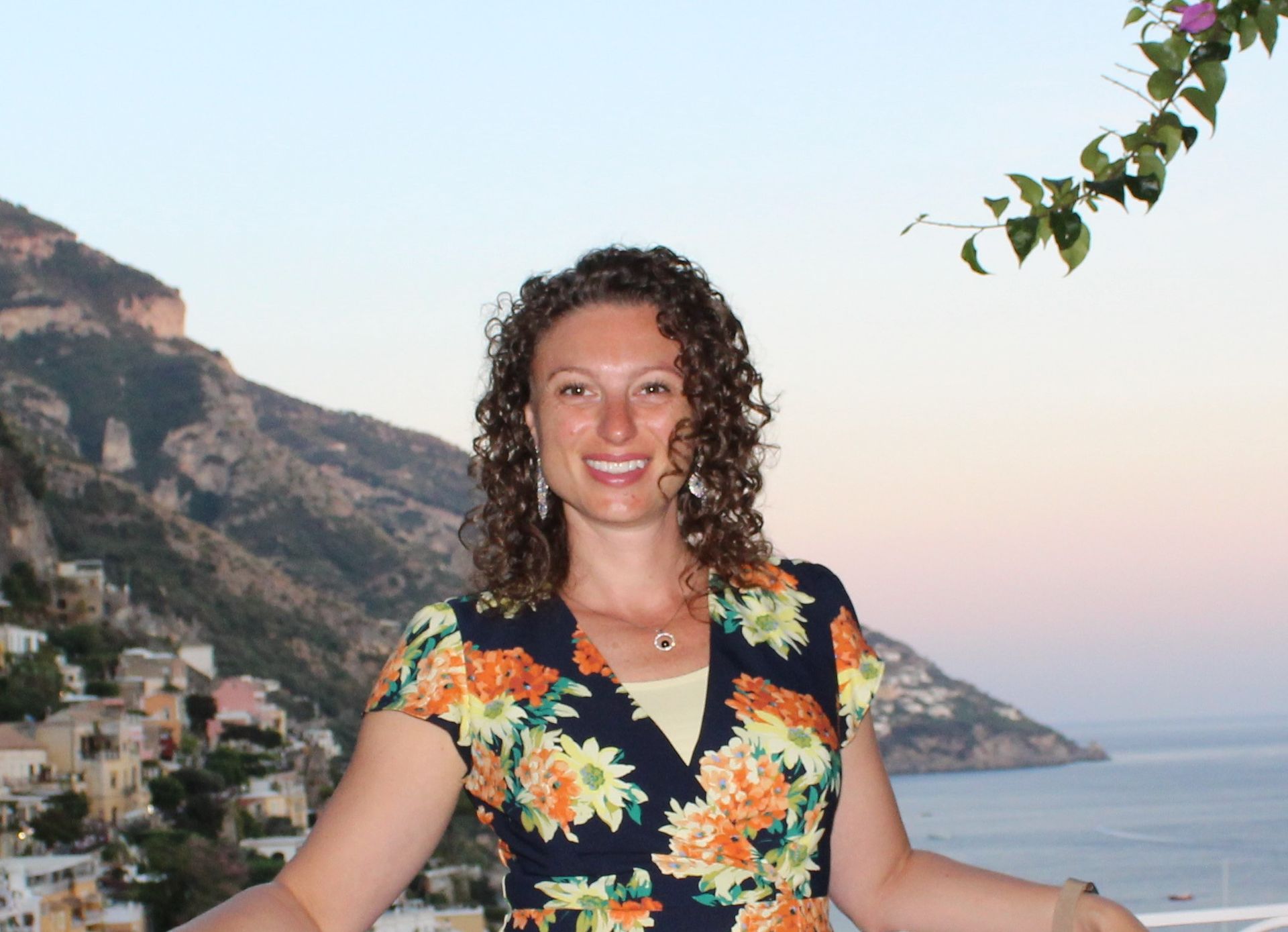 A woman in a floral dress stands in front of a mountain and ocean
