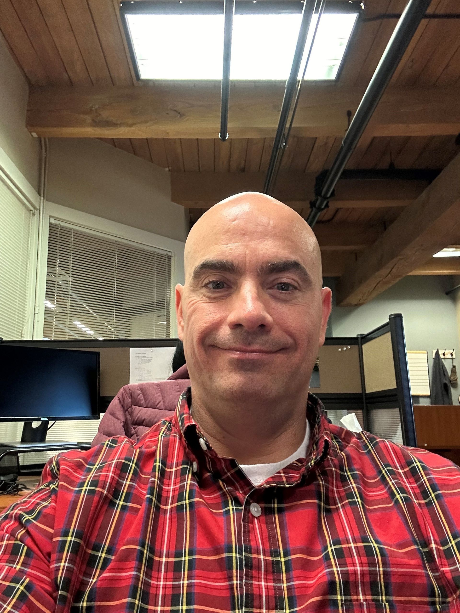 A bald man in a red plaid shirt is smiling in an office cubicle.