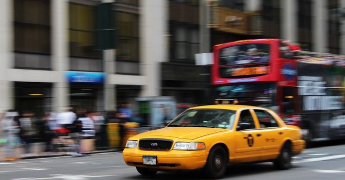 A yellow taxi is driving down a city street next to a red double decker bus.