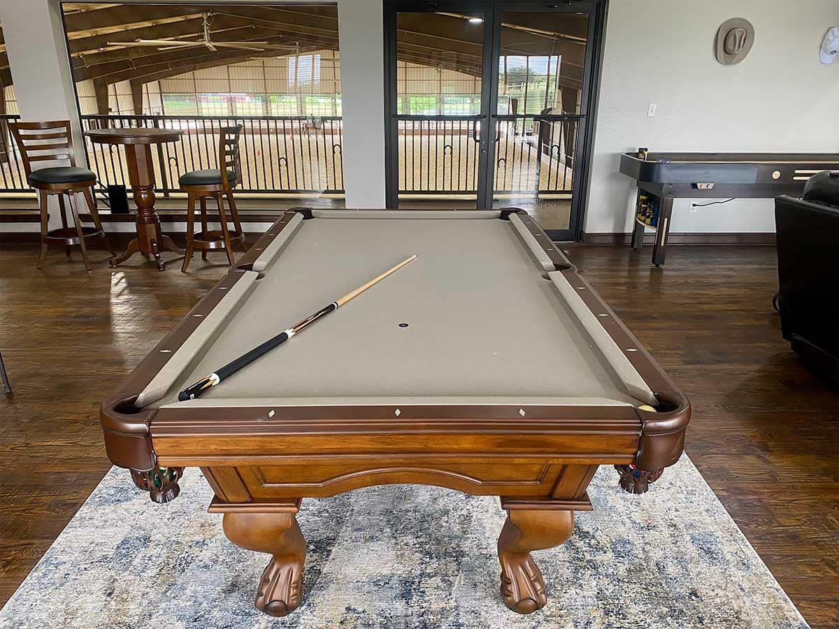 Pool table with a cue stick on a patterned rug in a room with a view of a barn.
